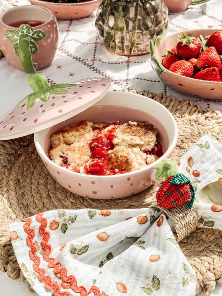 A bowl of strawberry cobbler sits on a woven placemat, surrounded by fresh strawberries, a strawberry-patterned napkin, and ceramic dishes decorated with strawberry motifs on a picnic table.