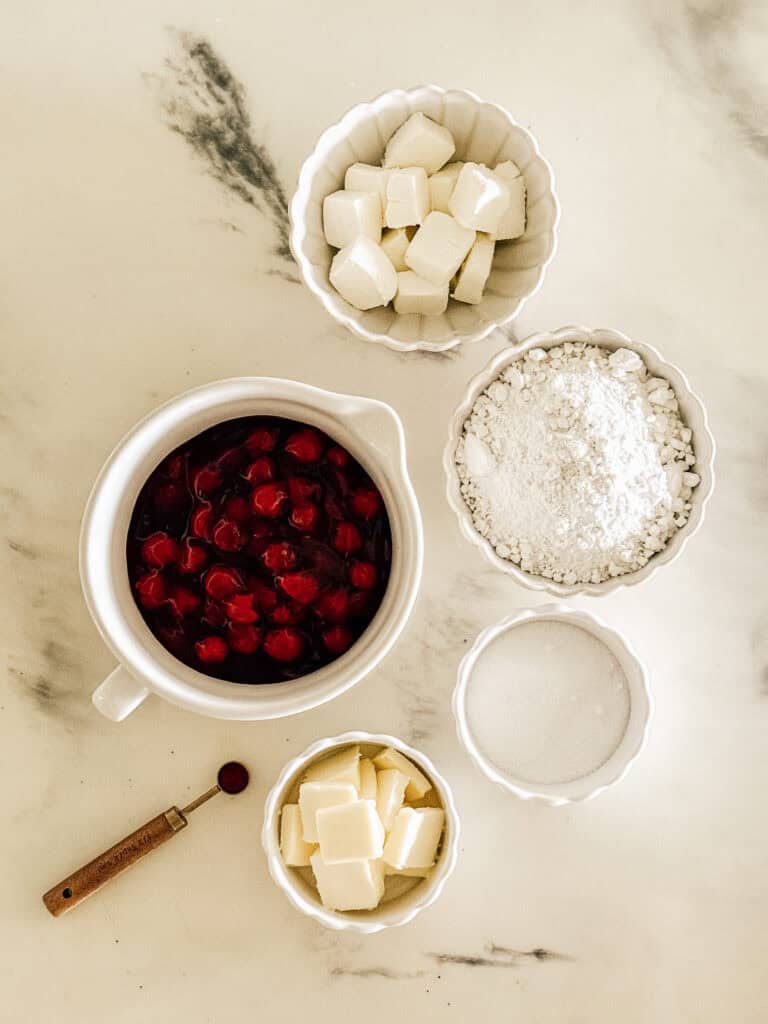 Five bowls hold cubed butter, cubed shortening, sugar, flour, and cherry pie filling on a marble countertop. A small measuring spoon lies beside the bowls.