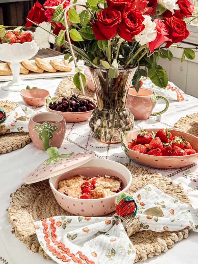 A table set with pink dishware, strawberries, cherries, a cobbler dessert, and a vase of red and white roses. Woven placemats, embroidered napkins, and a bright, summery atmosphere complete the scene.