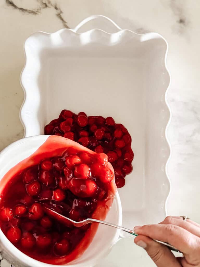 A hand spooning cherry pie filling from a bowl into a white scalloped baking dish on a marble counter.