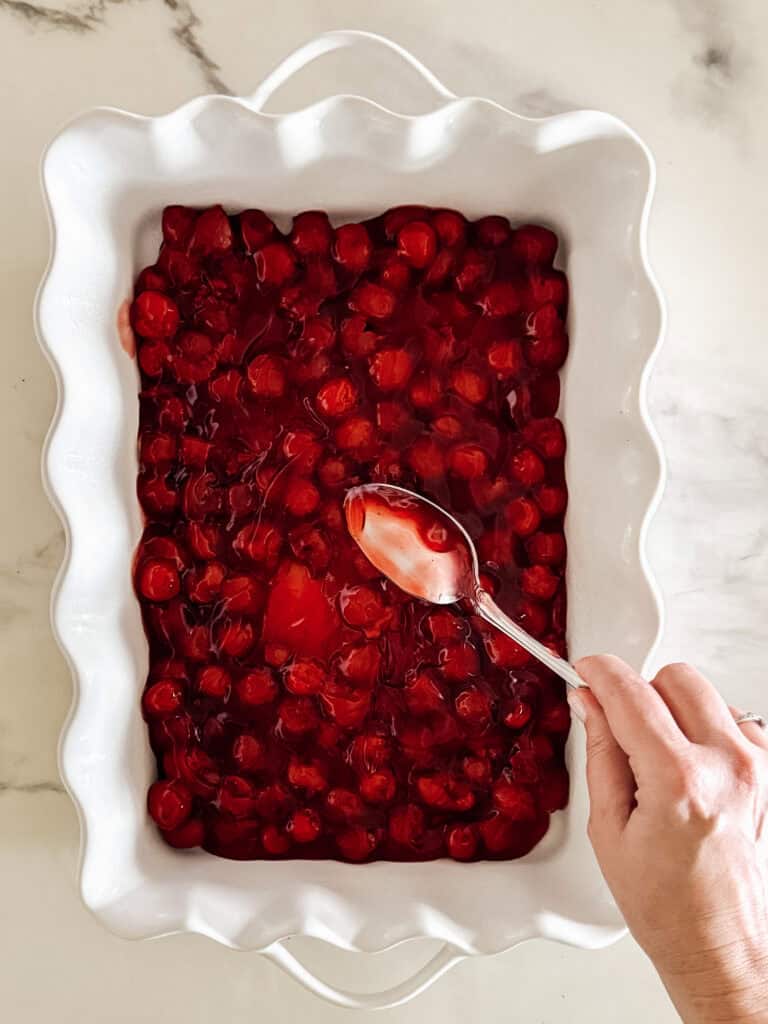 A hand holds a spoon scooping cherry pie filling from a white, wavy-edged baking dish on a marble surface.