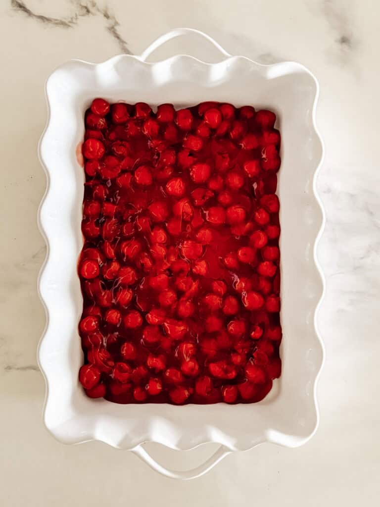 A white ceramic baking dish filled with bright red cherry pie filling sits on a light marble surface.