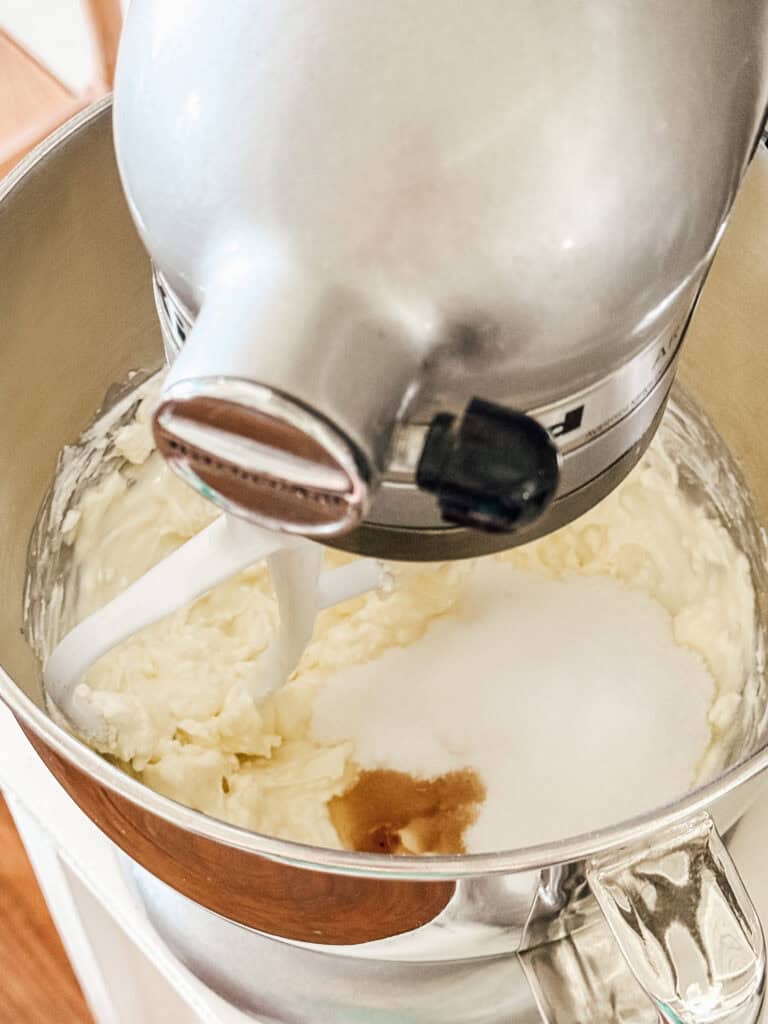 A stand mixer bowl contains softened butter, sugar, and vanilla extract, being mixed with a paddle attachment.