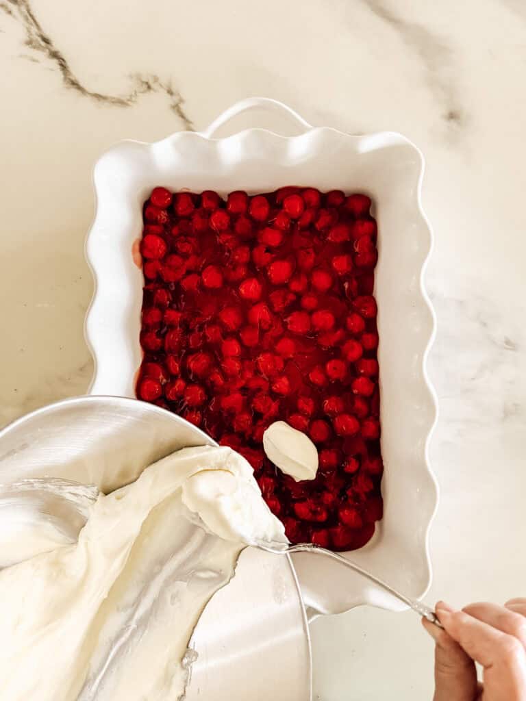 A hand scoops creamy batter from a metal bowl over a white dish filled with cherry pie filling on a marble countertop.