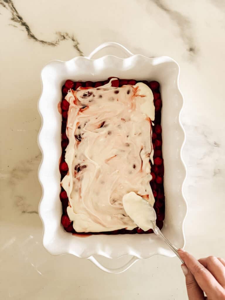 A hand spreads a creamy layer over a red fruit mixture in a white scalloped baking dish on a light marble countertop.