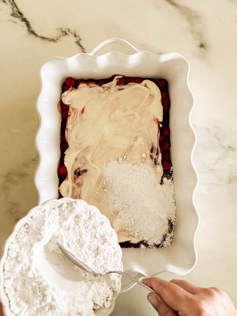 A hand holds a spoon over a white baking dish with cherries, creamy layer, and powdered mix, preparing to combine ingredients on a marble counter.